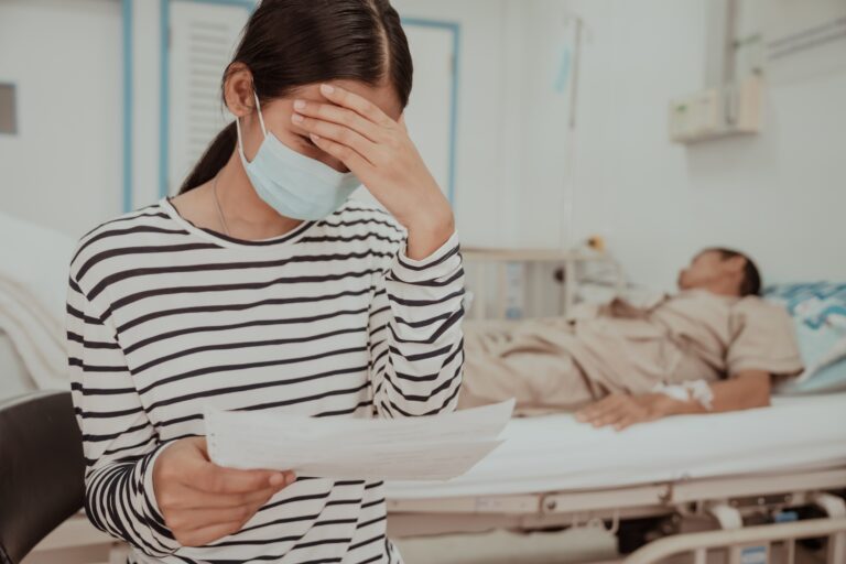 Woman sits by hospital bedside looking at medical bills